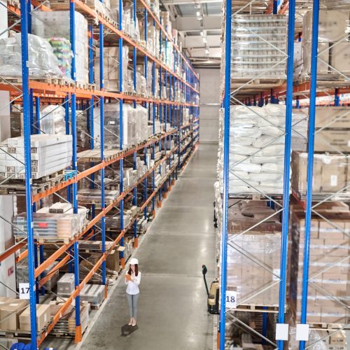 Large warehouse. Woman in white protective helmet with tablet standing in large industrial warehouse in aisle between high racks for goods
